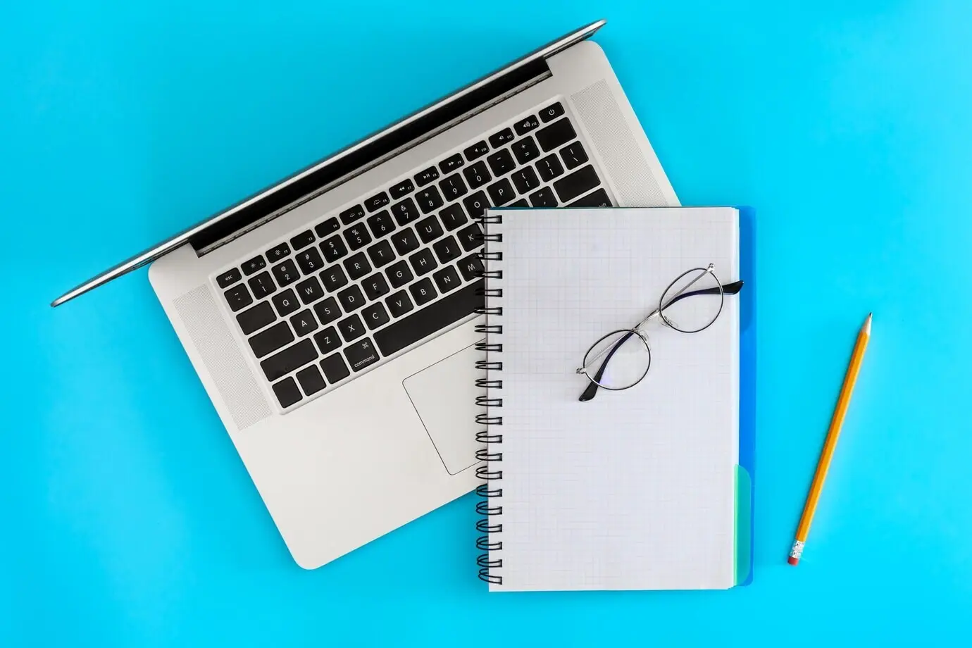 Draufsicht: Leeres Notizbuch, Laptop und Brille auf blauem Hintergrund.